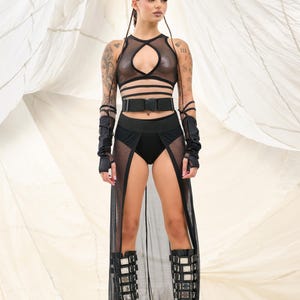 May include: A woman wearing a black mesh top with a cut-out design, a black belt, and a black mesh skirt with a slit. She is also wearing black boots with silver buckles.