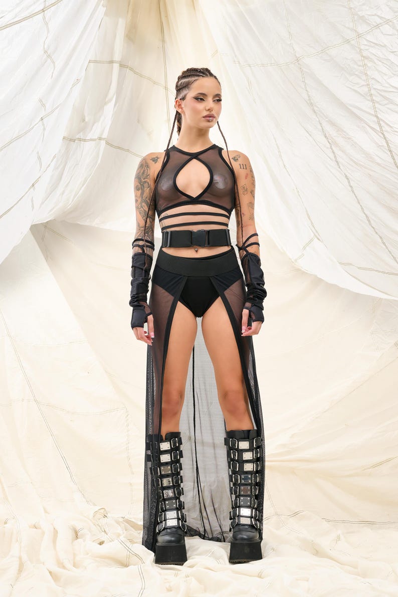 May include: A woman wearing a black mesh top with a cut-out design, a black belt, and a black mesh skirt with a slit. She is also wearing black boots with silver buckles.