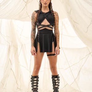 Pode incluir: Um conjunto preto de duas pe&ccedil;as com um top halter com detalhes cruzados e uma saia com fendas altas. O conjunto &eacute; combinado com botas de plataforma com v&aacute;rias fivelas. O fundo &eacute; um tecido neutro, branco sujo.