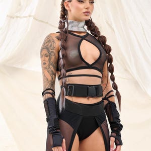 May include: A woman wearing a black mesh crop top with a cut-out design, a black belt with a buckle, and a black mesh skirt with a slit. She has long brown hair in braids and is wearing black fingerless gloves.