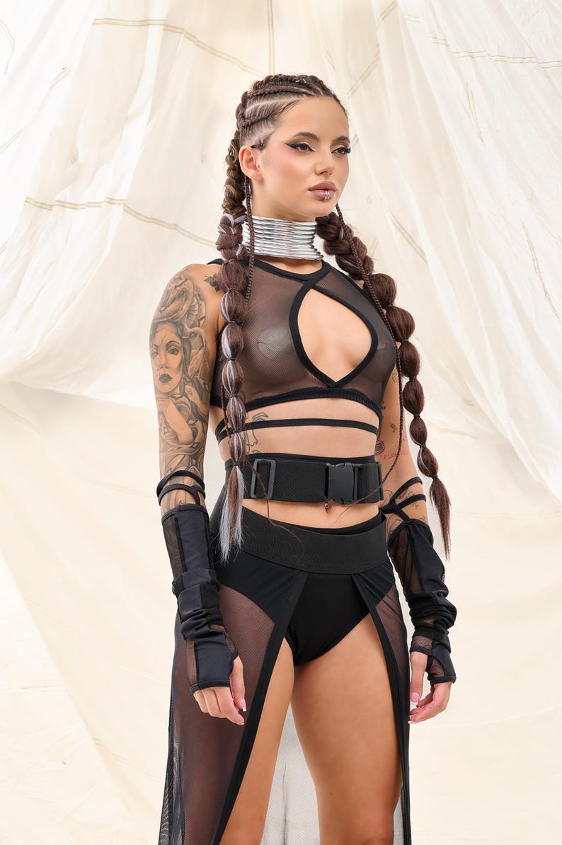 May include: A woman wearing a black mesh crop top with a cut-out design, a black belt with a buckle, and a black mesh skirt with a slit. She has long brown hair in braids and is wearing black fingerless gloves.