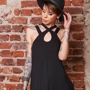 May include: A woman wearing a black sleeveless dress with a criss-cross neckline and a black fedora hat. She has short brown hair and tattoos on her arms.