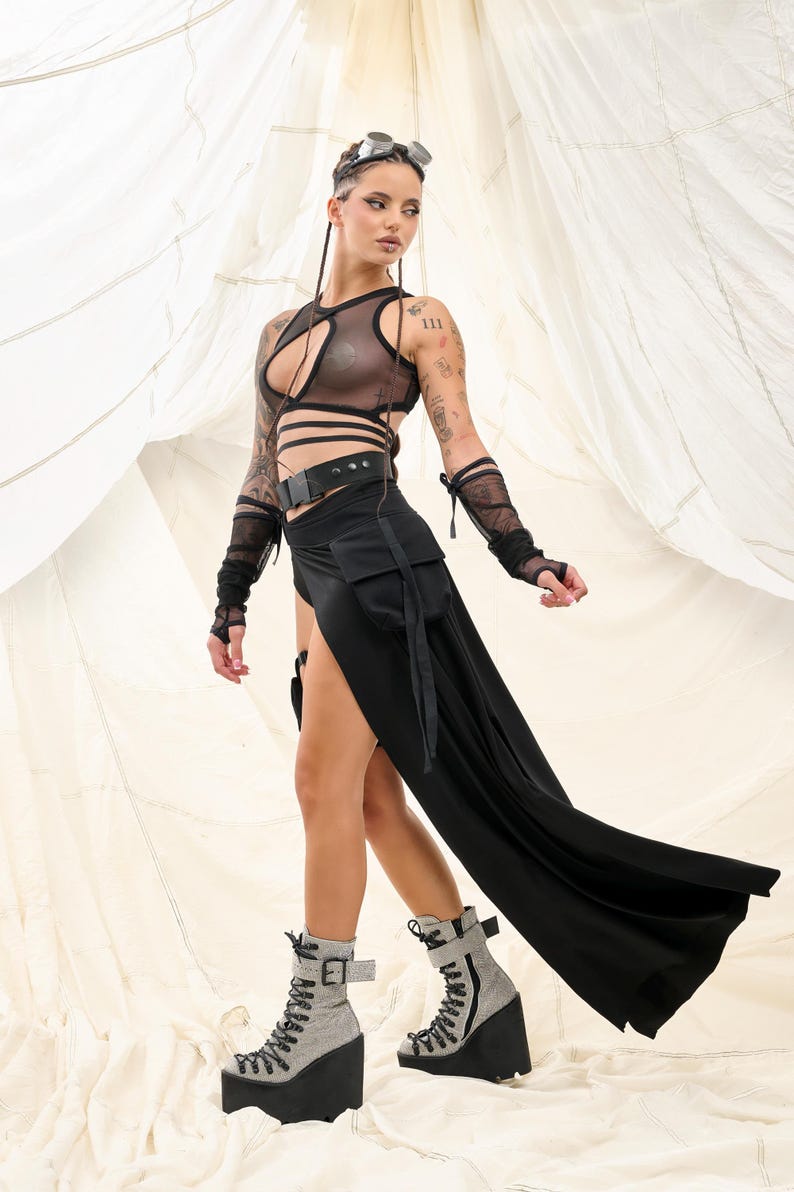 May include: A woman wearing a black mesh top with a black belt and a long black skirt with a side slit. She is wearing black lace gloves and grey platform boots with black laces.