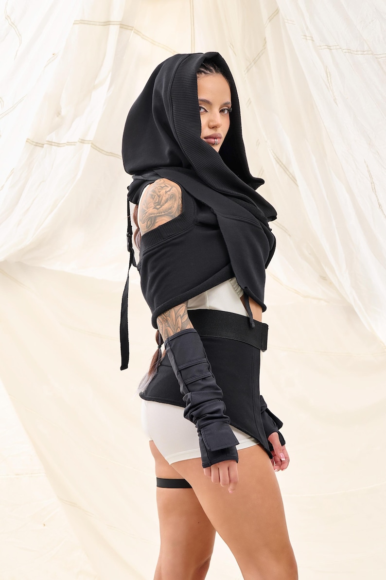 Op de afbeelding: Een zwart hooded top met een lange, vloeiende sjaal. De top wordt gedragen over een wit tanktop en een zwarte short. De persoon draagt zwarte vingerloze handschoenen.