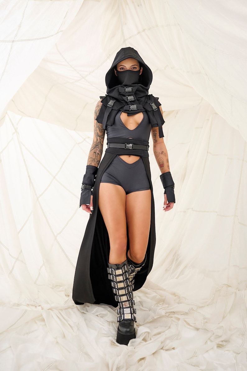 以下が含まれることがあります： 黒いフード付きの服を着て、顔にマスク、黒いバックル付きのベルト、黒いショートパンツを履いた人。その人は銀色のバックルが付いた黒いブーツを履いて、白い表面の上に立っている。