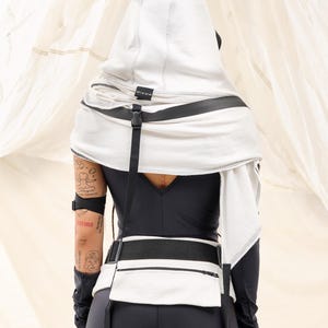 K&ouml;nnte beinhalten: Ein wei&szlig;es Kapuzenoberteil mit schwarzen Akzenten, darunter ein G&uuml;rtel mit einer Tasche und Riemen. Das Kleidungsst&uuml;ck hat ein minimalistisches Design mit sichtbaren N&auml;hten und einem Logo. Das Model tr&auml;gt schwarze Leggings und ein lang&auml;rmliges Oberteil.