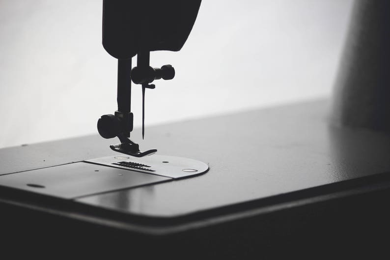 May include: Close-up of a sewing machine needle and foot, with the needle in the down position. The machine is black and the needle is silver.