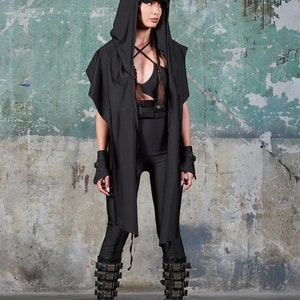 May include: A person wearing a black hooded top with a black belt and black boots. The person is standing in front of a green wall.
