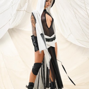 K&ouml;nnte beinhalten: Ein futuristisch anmutendes Outfit mit einer wei&szlig;en Kapuzenweste mit schwarzem Besatz, kombiniert mit einem durchsichtigen schwarzen Body und einem flie&szlig;enden schwarzen Rock. Das Ensemble umfasst schwarze Plateau-Stiefel und Kniesch&uuml;tzer, was einen k&uuml;hnen, avantgardistischen Look erzeugt.