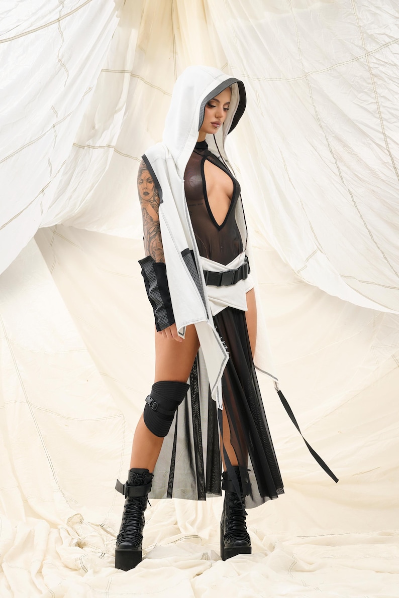 K&ouml;nnte beinhalten: Ein futuristisch anmutendes Outfit mit einer wei&szlig;en Kapuzenweste mit schwarzem Besatz, kombiniert mit einem durchsichtigen schwarzen Body und einem flie&szlig;enden schwarzen Rock. Das Ensemble umfasst schwarze Plateau-Stiefel und Kniesch&uuml;tzer, was einen k&uuml;hnen, avantgardistischen Look erzeugt.