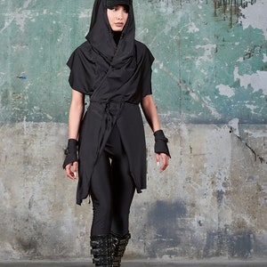 May include: A person wearing a black hooded top with a long, flowing design, black trousers, and black boots with buckles. The person is standing in front of a green and grey textured wall.