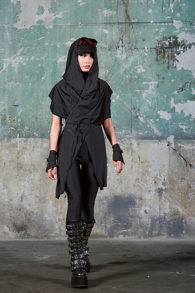 May include: A person wearing a black hooded top with a long, flowing design, black trousers, and black boots with buckles. The person is standing in front of a green and grey textured wall.