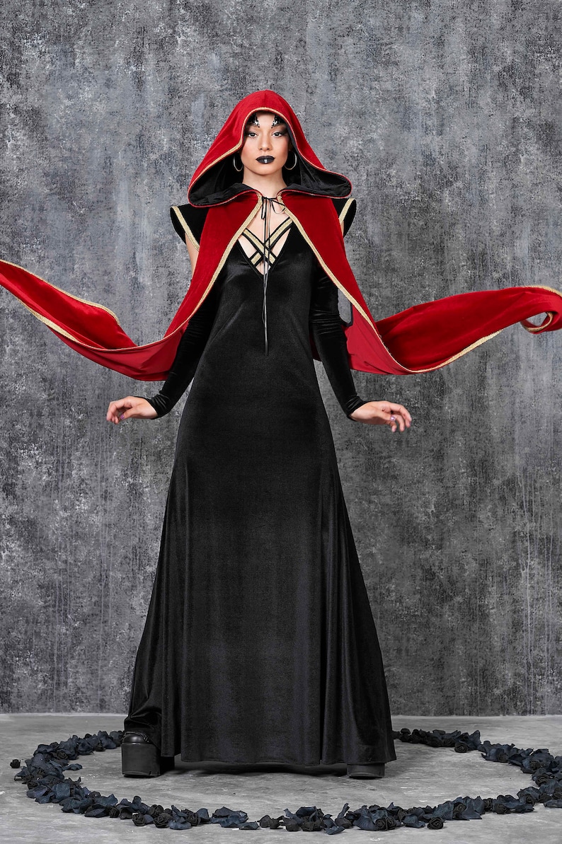May include: A black velvet gown with long sleeves and a flowing skirt. A red hooded cape with gold trim is draped over the shoulders. The cape's fabric is flowing. The outfit is suitable for a costume or gothic-themed event.