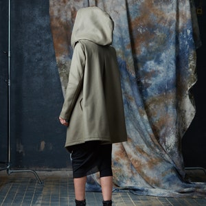 Puede incluir: Una chaqueta de color marr&oacute;n claro con capucha y un dise&ntilde;o largo y fluido. La chaqueta est&aacute; hecha de una tela suave y texturizada y tiene un corte holgado. La modelo lleva la chaqueta con una falda negra y botas negras.
