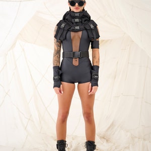 以下が含まれることがあります： 黒いボディスーツ、メッシュパネル、黒いベルト、バックル付きの黒いハーネスを着用した女性。黒い手袋、黒いブーツ、サングラスも着用している。