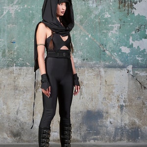 May include: A person wearing a black hooded top, black trousers, and black platform boots with buckles. The person is standing in front of a green and grey textured wall.