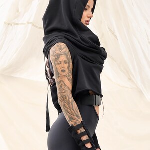 Op de afbeelding: Een zwart hoodie met een lange, vloeiende cowl neck. De top is gecropped en heeft een riem in de taille. Het model draagt ​​zwarte leggings en heeft een tatoeage op haar arm.