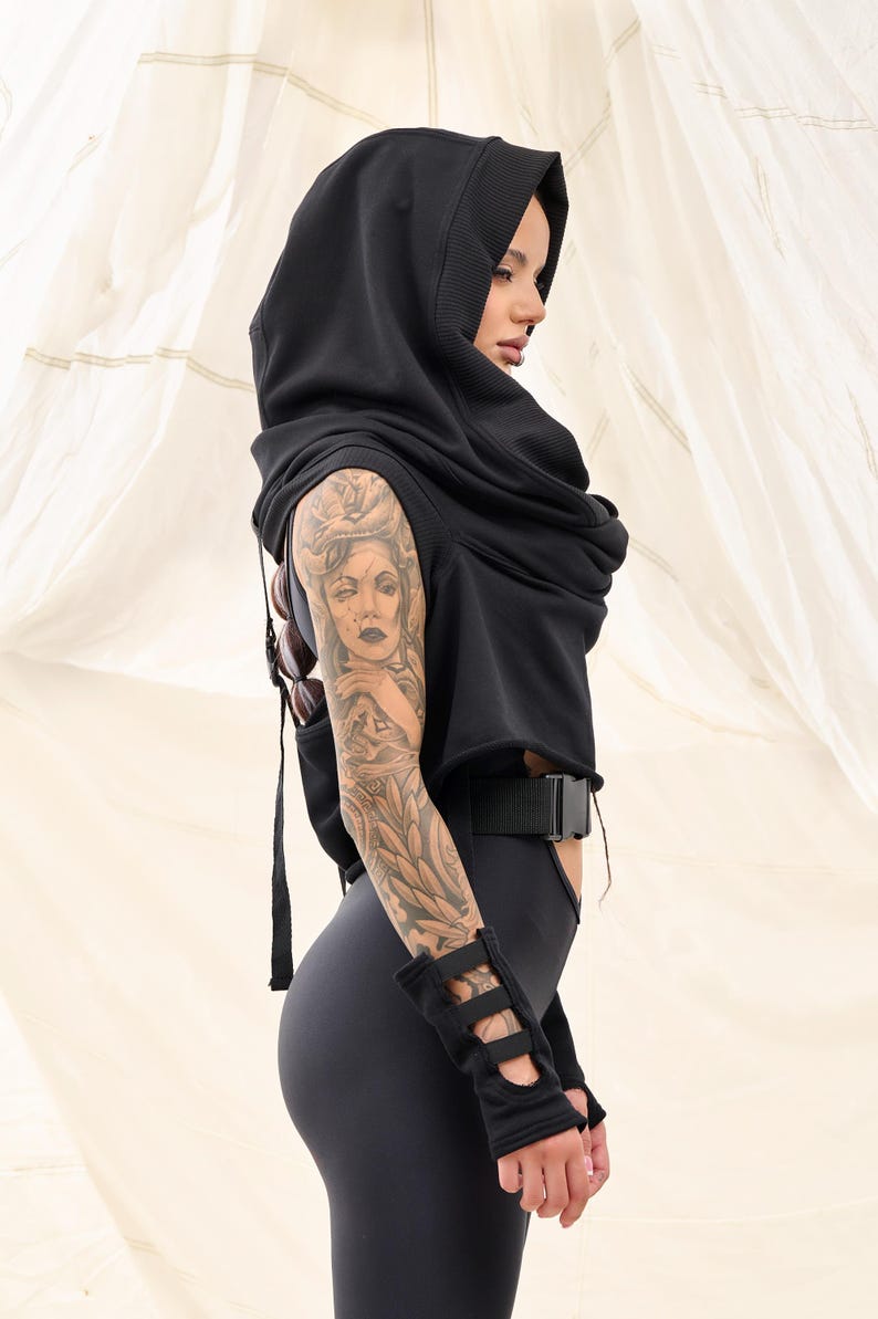 Op de afbeelding: Een zwart hoodie met een lange, vloeiende cowl neck. De top is gecropped en heeft een riem in de taille. Het model draagt ​​zwarte leggings en heeft een tatoeage op haar arm.
