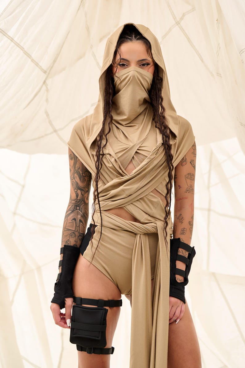 May include: A tan-colored hooded bodysuit with a face covering and wrap-around design. The outfit includes black wrist cuffs and a thigh-mounted pouch. The model has tattoos on their arms. The background is a light-colored fabric.