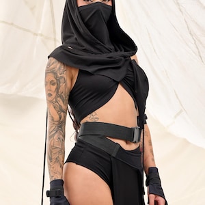 Pu&ograve; includere: Una persona che indossa un top nero con cappuccio e maschera facciale, un top corto nero, una cintura nera e pantaloncini neri. La persona ha un tatuaggio sul braccio.