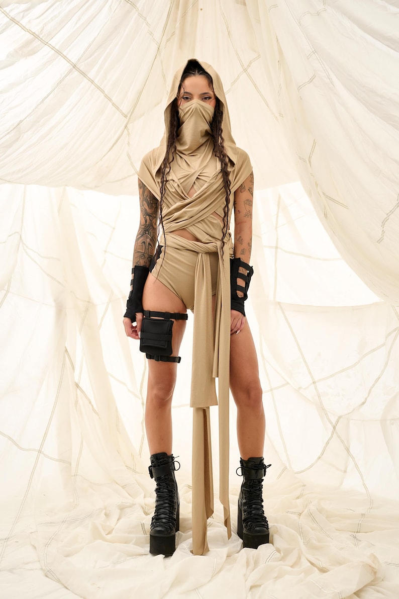 May include: A tan-colored hooded outfit with a face covering, wrap-around top, and high-waisted shorts. The outfit includes black arm cuffs, a leg holster, and platform boots. The background is a light-colored fabric.