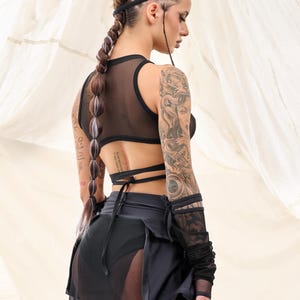 May include: A woman wearing a black mesh crop top with a black skirt and a sheer black overlay. She has a large tattoo on her right arm and a braided hairstyle with white accents.