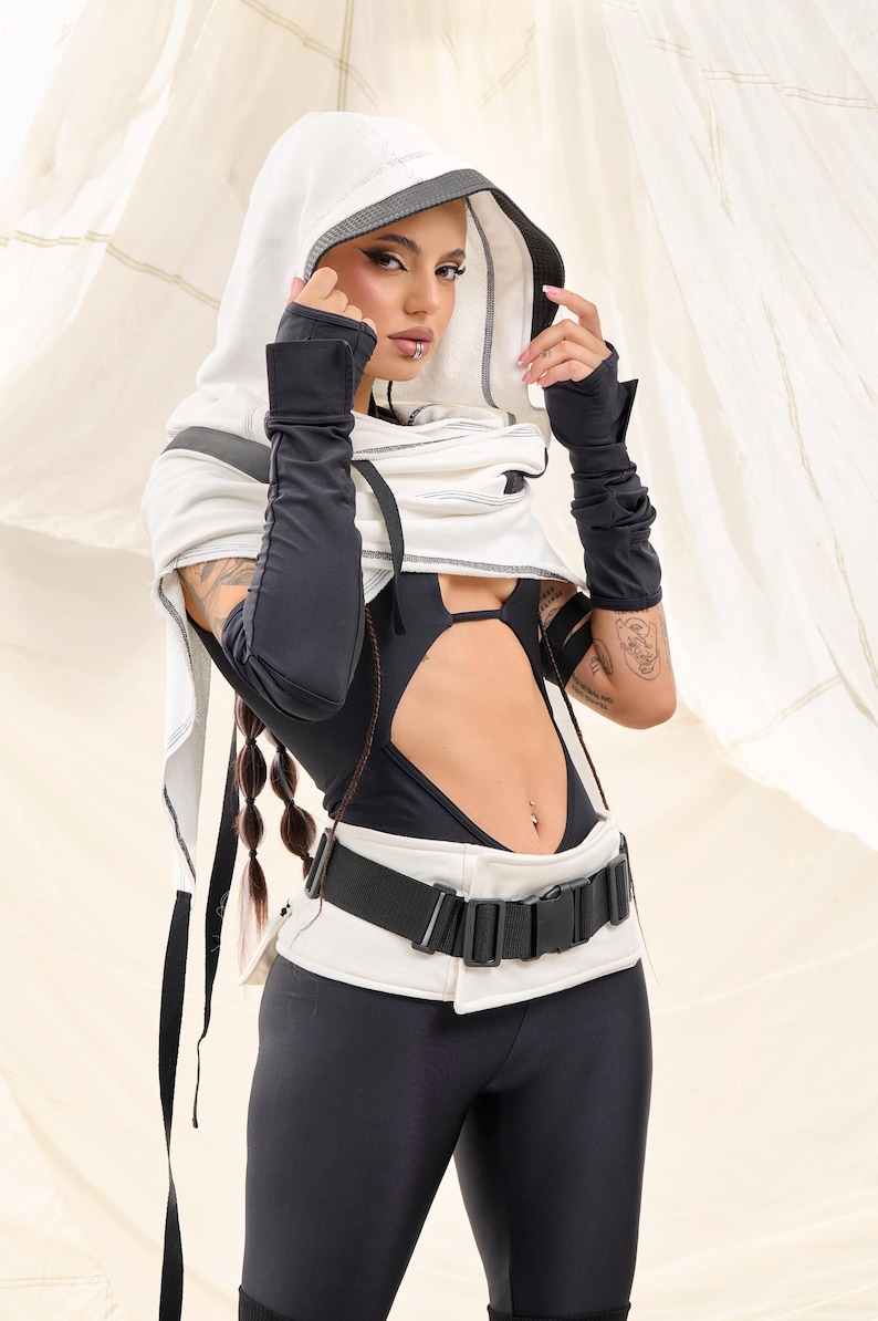 K&ouml;nnte beinhalten: Ein futuristisch anmutendes Outfit in Schwarz und Wei&szlig;. Das Ensemble umfasst ein Kapuzentop, lange Handschuhe, einen Body mit Ausschnitten und Leggings. Ein breiter G&uuml;rtel mit Schnalle betont die Taille. Das Outfit hat eine moderne, kantige &Auml;sthetik.