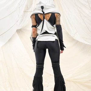 K&ouml;nnte beinhalten: Ein futuristisches Outfit mit einem wei&szlig;en Kapuzenoberteil mit schwarzen Akzenten, einer passenden Weste und schwarzen Leggings. Das Ensemble umfasst lange schwarze Handschuhe und eine ausgestellte Hose mit Cargotaschen. Das Outfit ist vor einem wei&szlig;en Hintergrund.