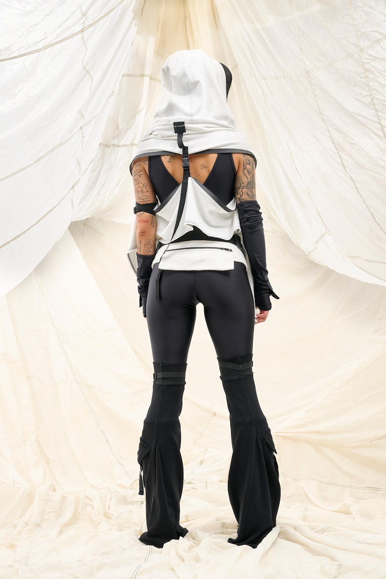 K&ouml;nnte beinhalten: Ein futuristisches Outfit mit einem wei&szlig;en Kapuzenoberteil mit schwarzen Akzenten, einer passenden Weste und schwarzen Leggings. Das Ensemble umfasst lange schwarze Handschuhe und eine ausgestellte Hose mit Cargotaschen. Das Outfit ist vor einem wei&szlig;en Hintergrund.