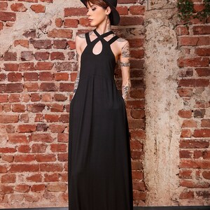 May include: A woman wearing a black maxi dress with a criss-cross halter neckline and a black hat. She is standing in front of a brick wall.