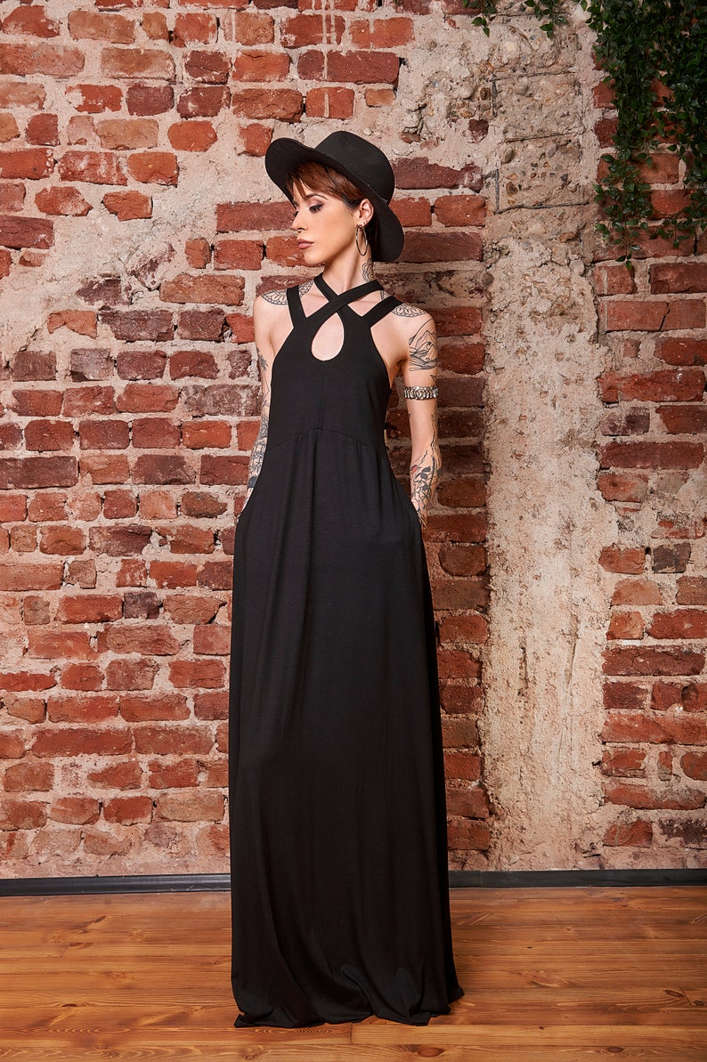May include: A woman wearing a black maxi dress with a criss-cross halter neckline and a black hat. She is standing in front of a brick wall.