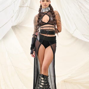 May include: A woman wearing a black mesh skirt with a high slit, a brown leather halter top with black straps, a black belt with a silver buckle, and black platform boots with silver buckles. She is standing in front of a white fabric backdrop.