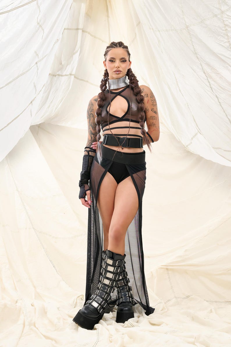 May include: A woman wearing a black mesh skirt with a high slit, a brown leather halter top with black straps, a black belt with a silver buckle, and black platform boots with silver buckles. She is standing in front of a white fabric backdrop.
