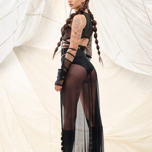 May include: A woman wearing a black mesh skirt and a black crop top with straps. She has long brown hair in braids and is wearing black boots.