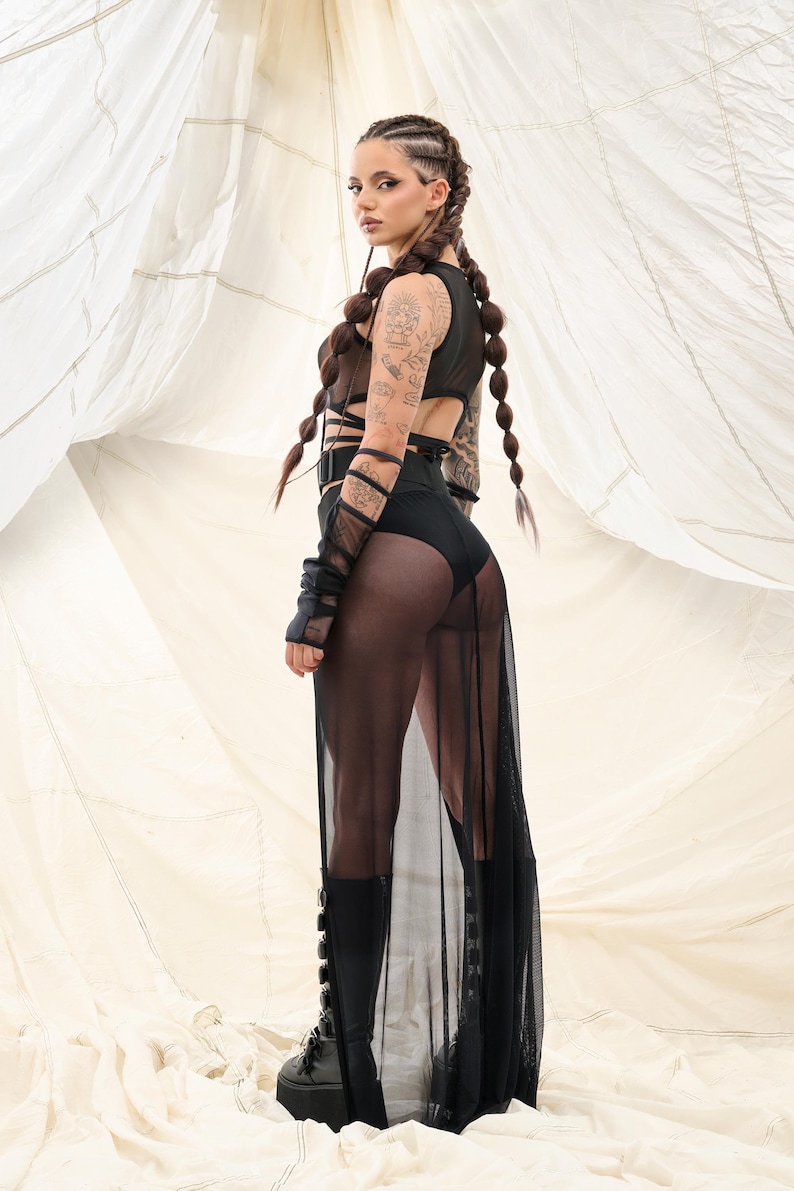 May include: A woman wearing a black mesh skirt and a black crop top with straps. She has long brown hair in braids and is wearing black boots.