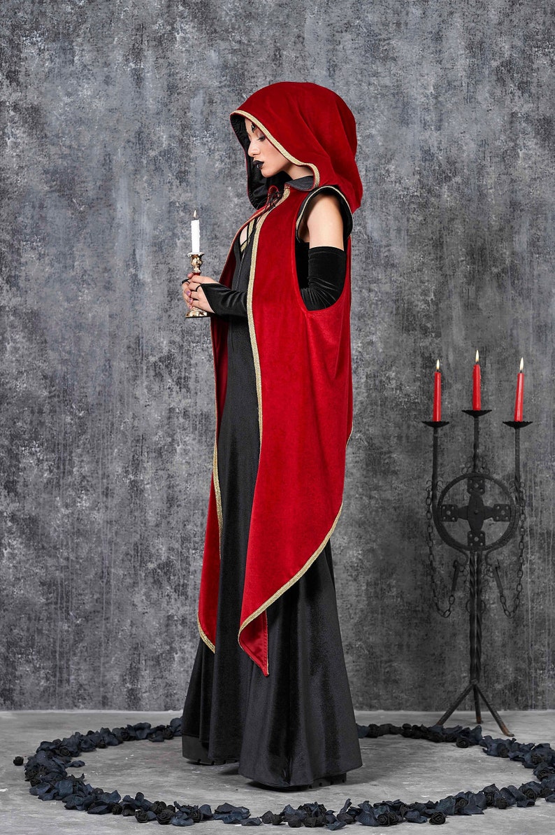 May include: A black dress is paired with a red hooded cape with gold trim. The person is holding a candle in a gold candlestick. A wrought iron candelabra with three red candles is in the background. The outfit is gothic-inspired.