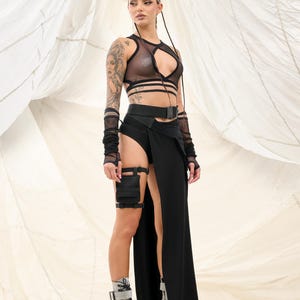 May include: A woman wearing a black mesh crop top with a black belt and a long black skirt with a slit. She is also wearing black fingerless gloves and black boots.