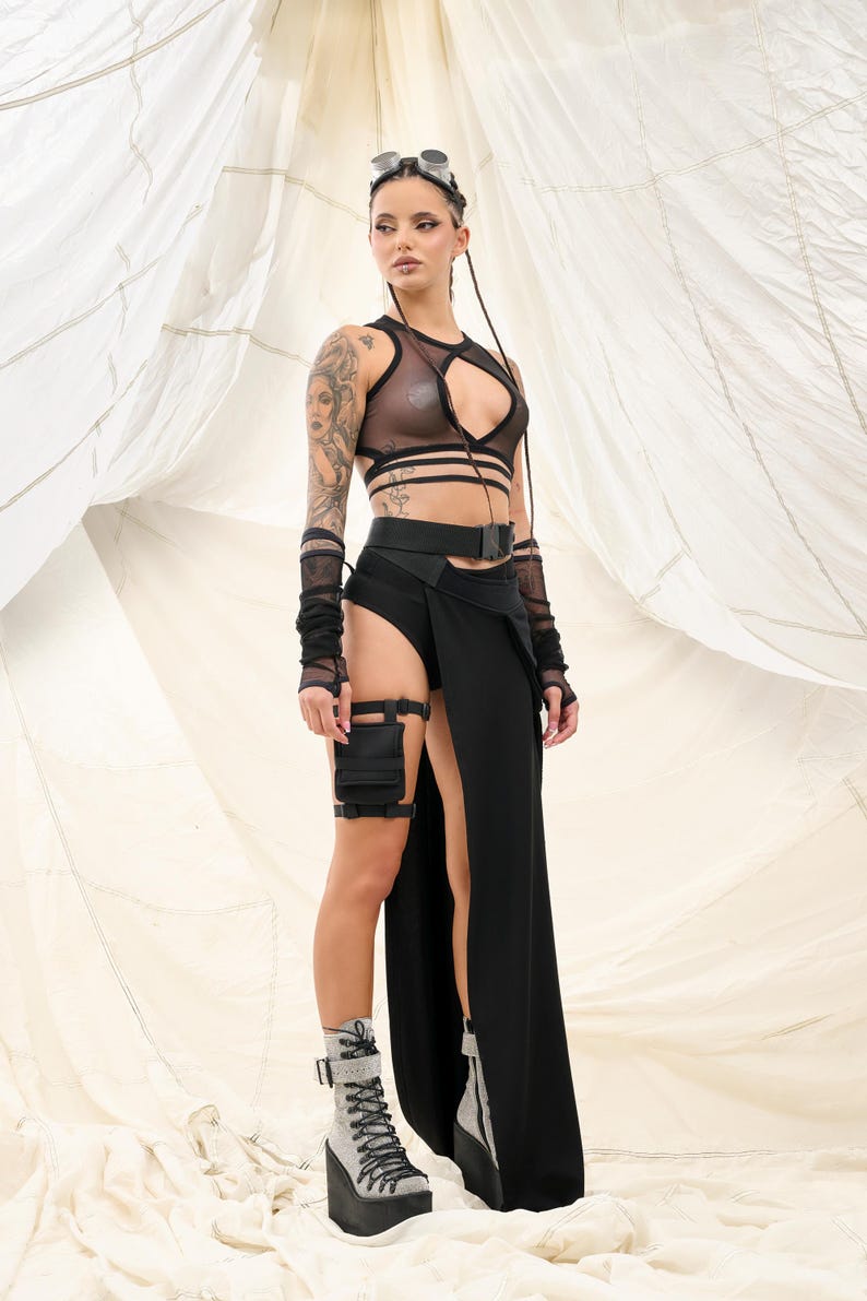 May include: A woman wearing a black mesh crop top with a black belt and a long black skirt with a slit. She is also wearing black fingerless gloves and black boots.
