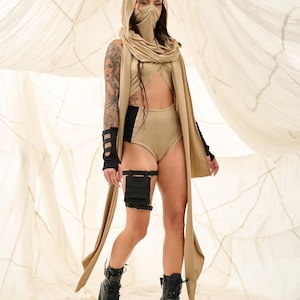 May include: A tan-colored hooded outfit with a matching face covering and long scarf. The outfit includes a high-waisted bottom, a top with a cutout, and black arm and leg accessories. Black platform boots complete the look.