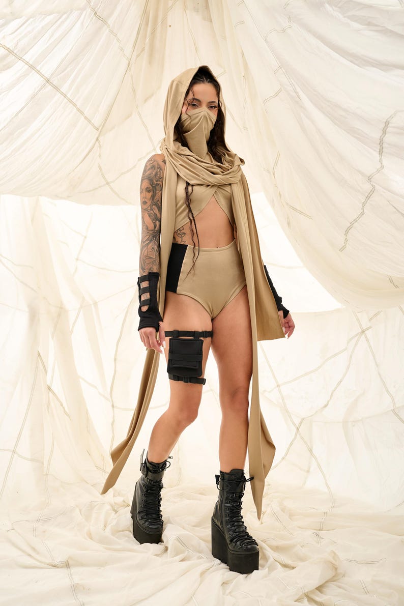 May include: A tan-colored hooded outfit with a matching face covering and long scarf. The outfit includes a high-waisted bottom, a top with a cutout, and black arm and leg accessories. Black platform boots complete the look.