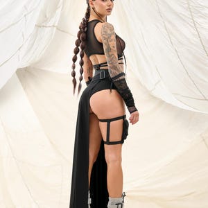 May include: A woman wearing a black mesh top with a black skirt and thigh-high garters. She is wearing silver platform boots and has her hair in braids. She is standing in front of a white fabric background.