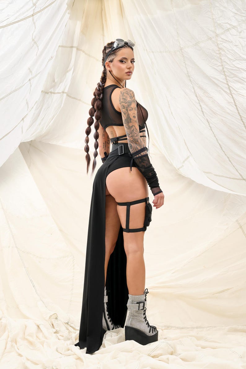 May include: A woman wearing a black mesh top with a black skirt and thigh-high garters. She is wearing silver platform boots and has her hair in braids. She is standing in front of a white fabric background.