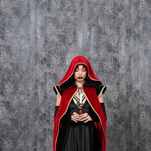 May include: A person wearing a red velvet hooded cloak with gold trim over a black dress, holding a candle. The background is a textured gray wall. The outfit is part of a gothic or fantasy theme.
