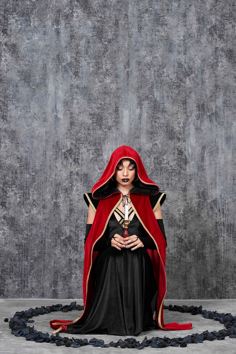 May include: A person wearing a red velvet hooded cloak with gold trim over a black dress, holding a candle. The background is a textured gray wall. The outfit is part of a gothic or fantasy theme.
