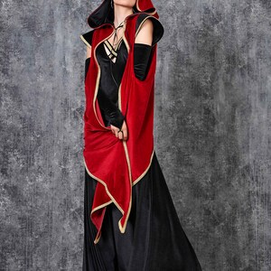May include: A black velvet dress with a red hooded cape trimmed in gold. The cape has long sleeves and a draped front. The outfit is worn with black platform boots. The background is a textured gray wall.
