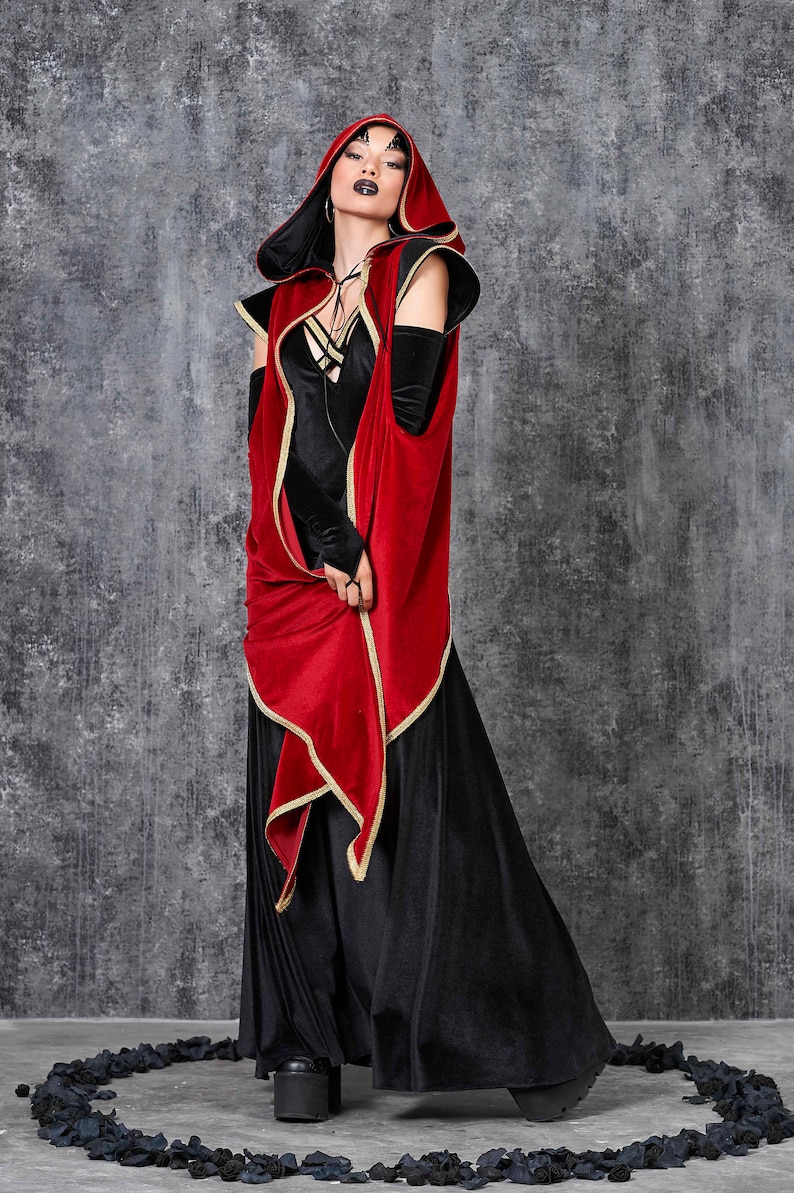 May include: A black velvet dress with a red hooded cape trimmed in gold. The cape has long sleeves and a draped front. The outfit is worn with black platform boots. The background is a textured gray wall.