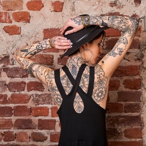 May include: A woman wearing a black hat and a black sleeveless dress with straps that cross at the back. She has tattoos on her arms, back, and neck.