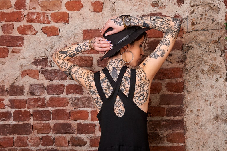 May include: A woman wearing a black hat and a black sleeveless dress with straps that cross at the back. She has tattoos on her arms, back, and neck.