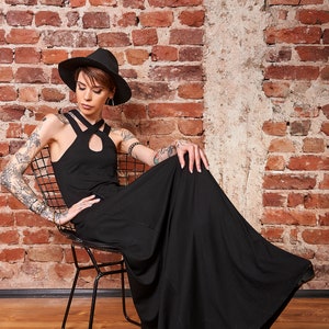 May include: A woman wearing a black maxi dress and a black fedora hat sits on a black metal chair. She has tattoos on her arms and is looking down. The background is a brick wall.