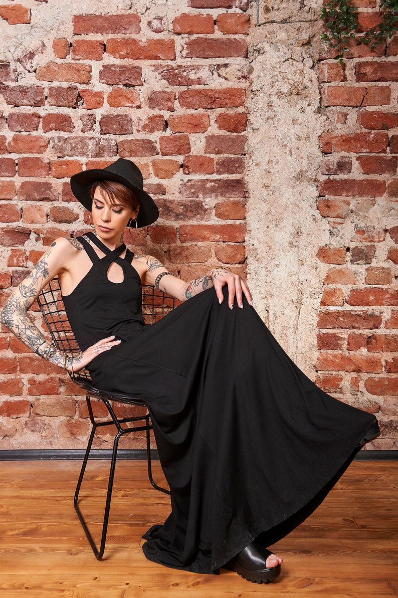May include: A woman wearing a black maxi dress and a black fedora hat sits on a black metal chair. She has tattoos on her arms and is looking down. The background is a brick wall.